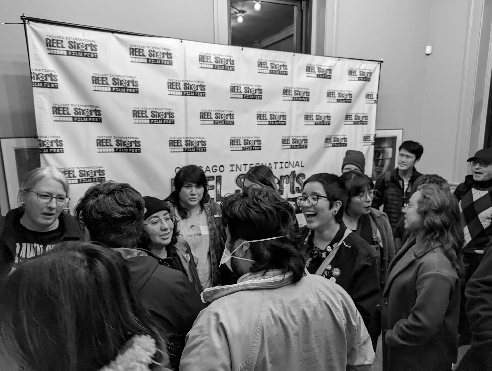 A group of people smiling and talking in front of a banner at an event. Black and white photo.