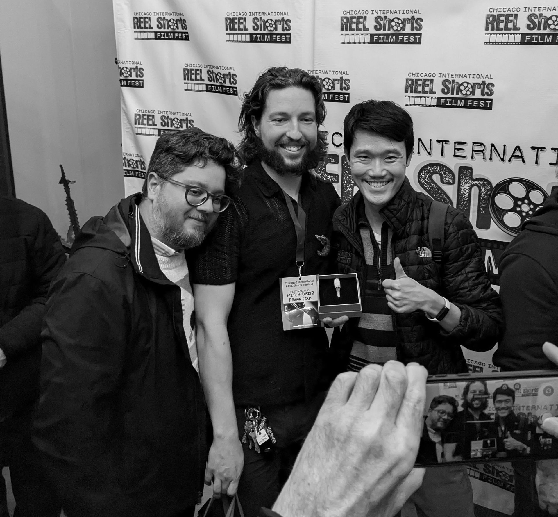 Three men smiling, posing for a photo at Reel Shorts Film Fest. One holds a booklet, one thumbs up.
