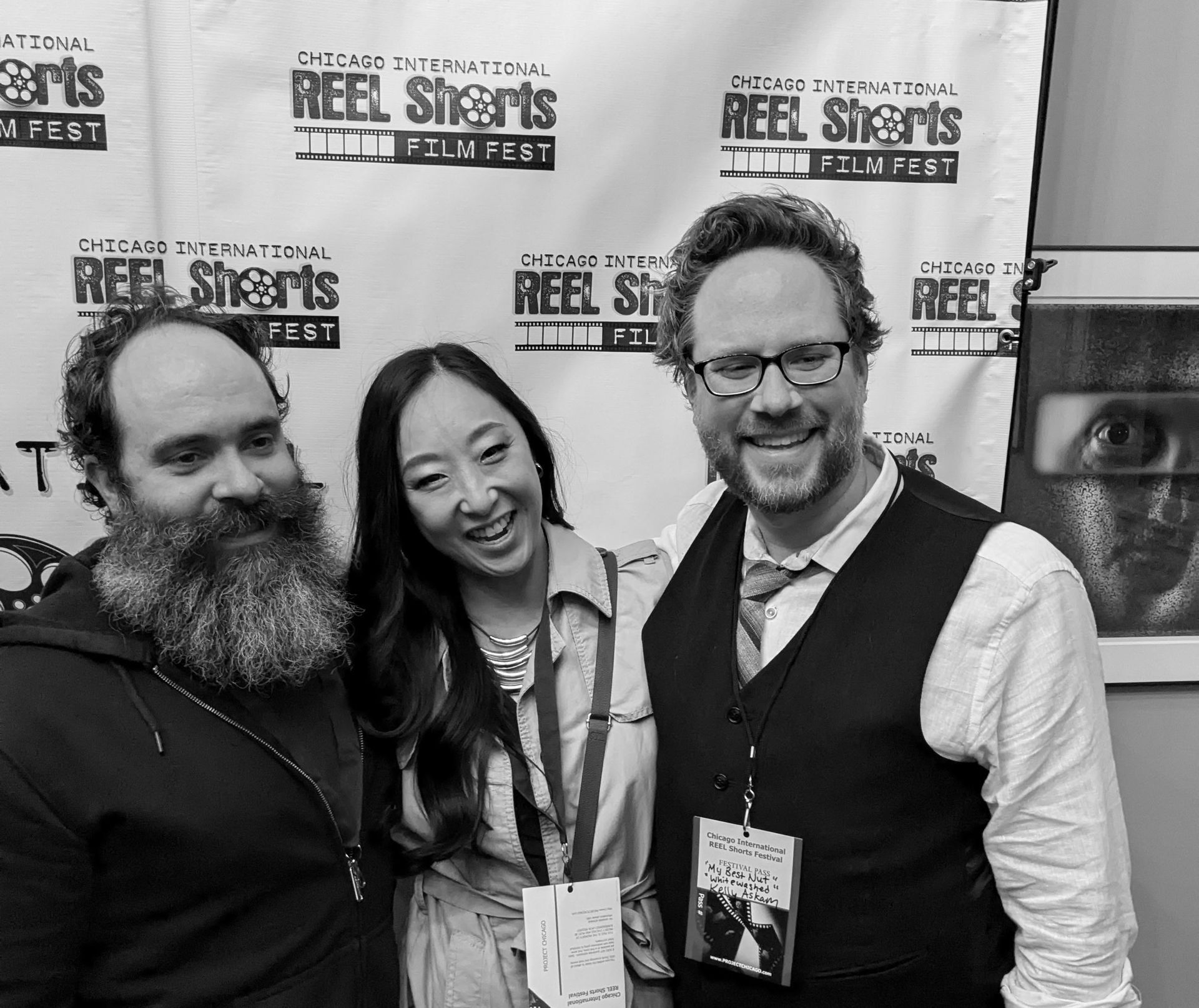 Three people smiling at a film festival. Man with beard, woman, man with glasses. Festival backdrop.