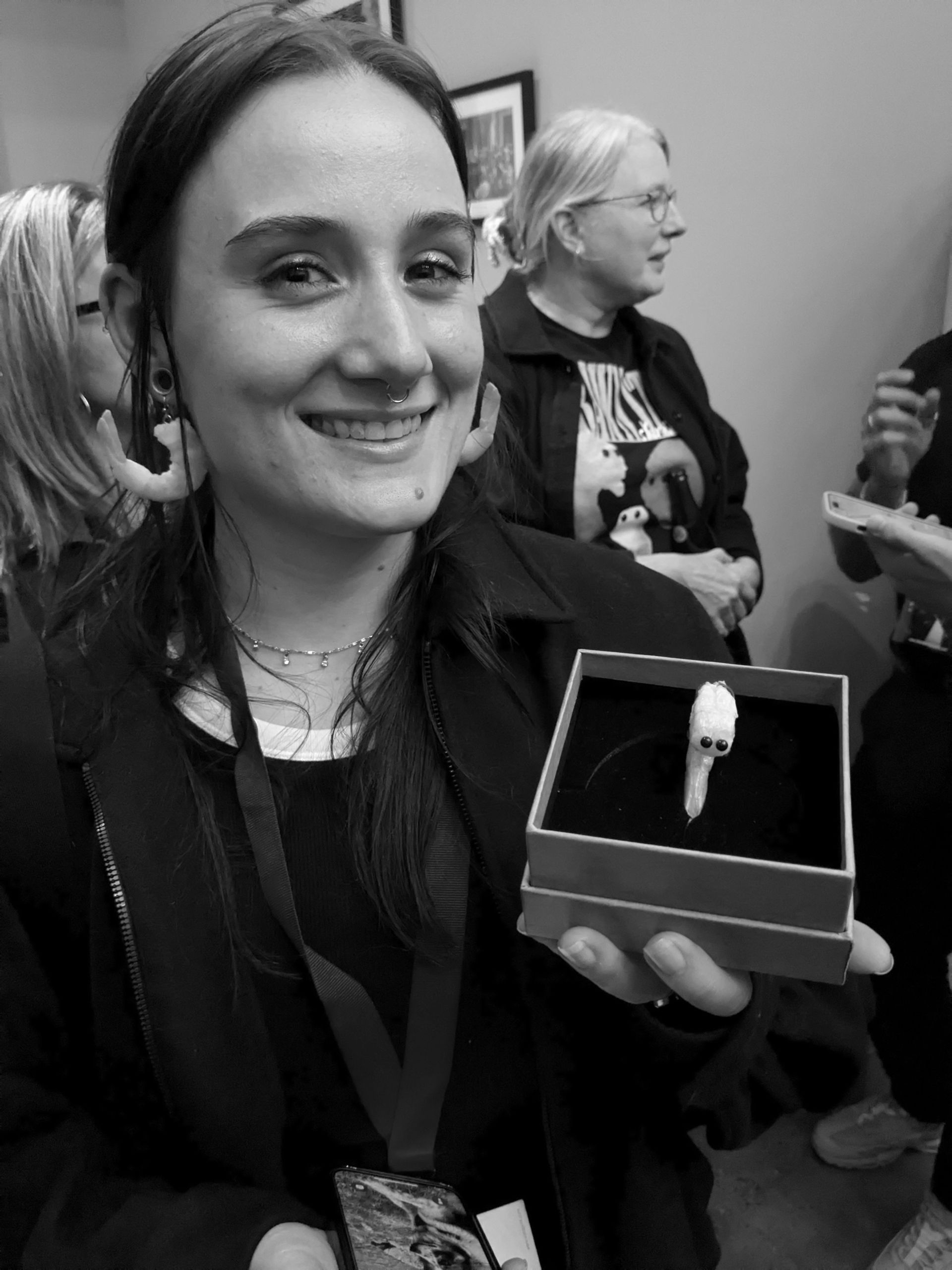 Woman smiles, holding a small box with a white object. People in the background. Black and white photo.