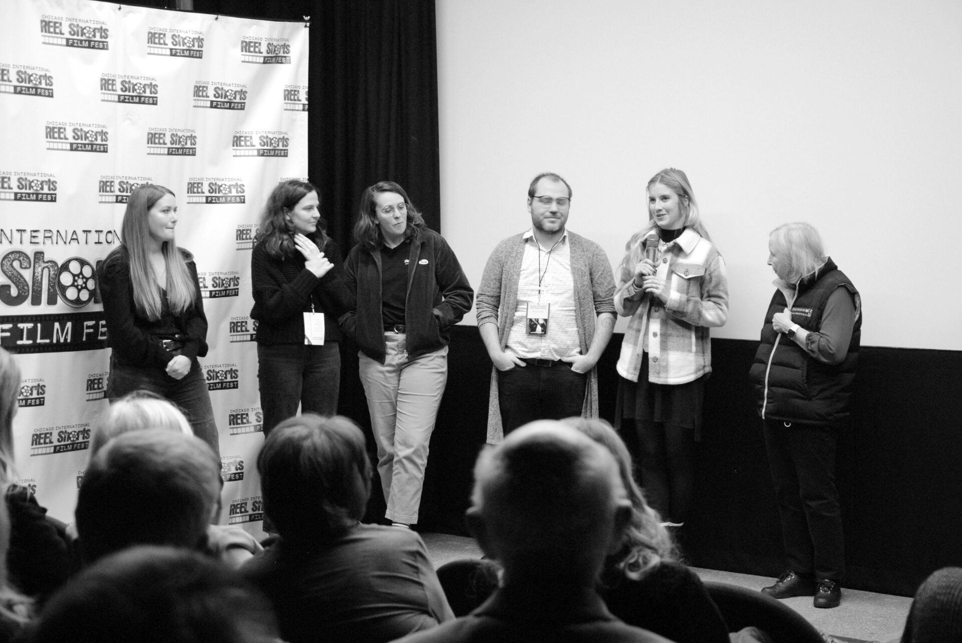 Panel of six people on stage, facing an audience. They are at a film festival, a blank screen behind them.
