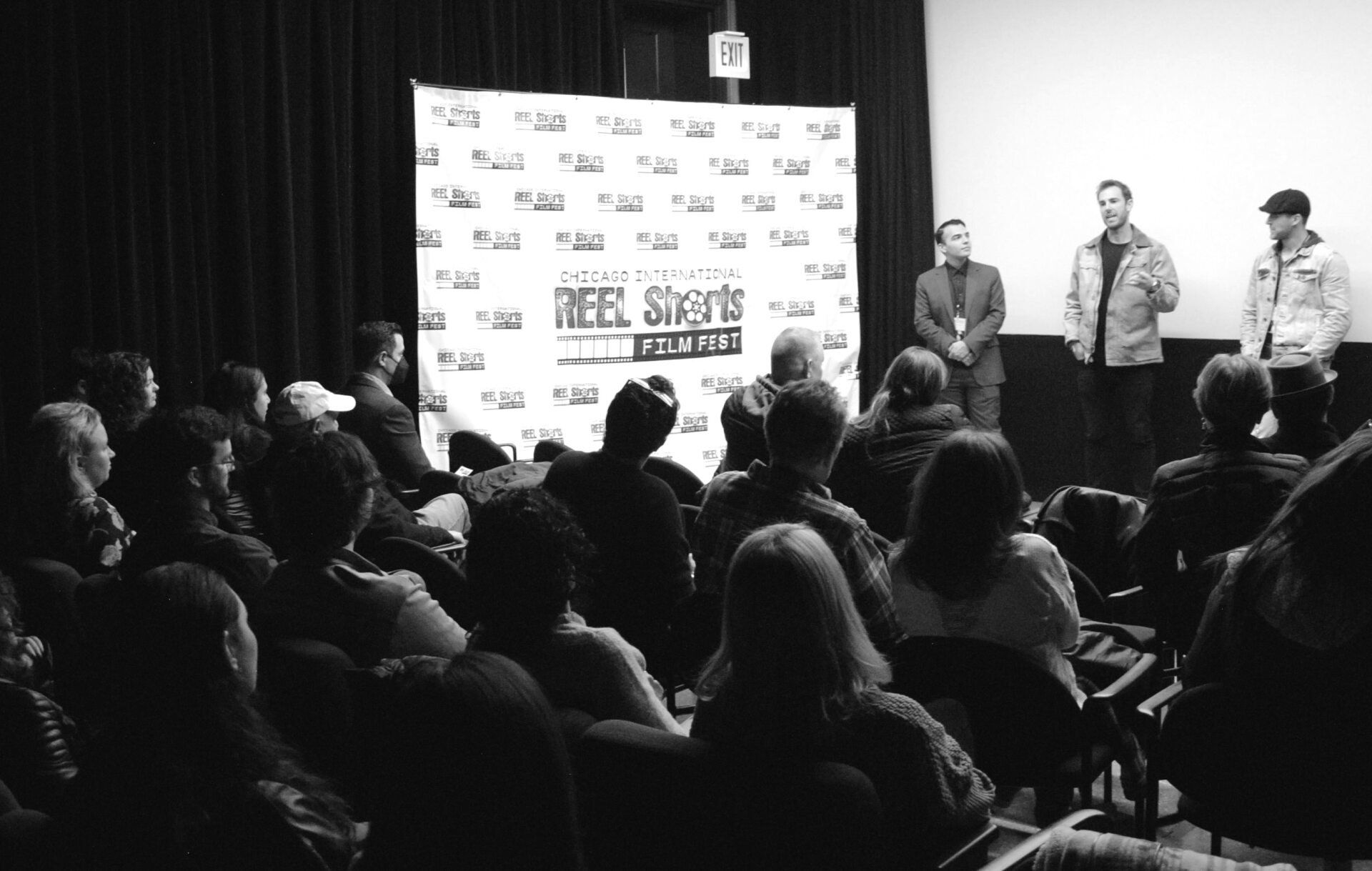 Audience watches a panel discussion at a film festival. Three men stand on stage, black and white photo.