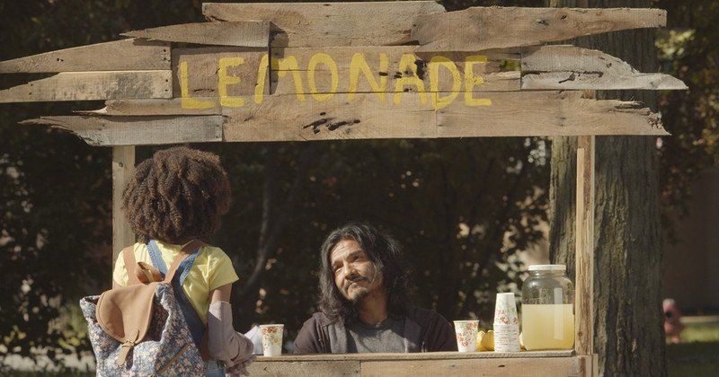 A person selling lemonade from a wooden stand to a young child. Sunny day; park setting.