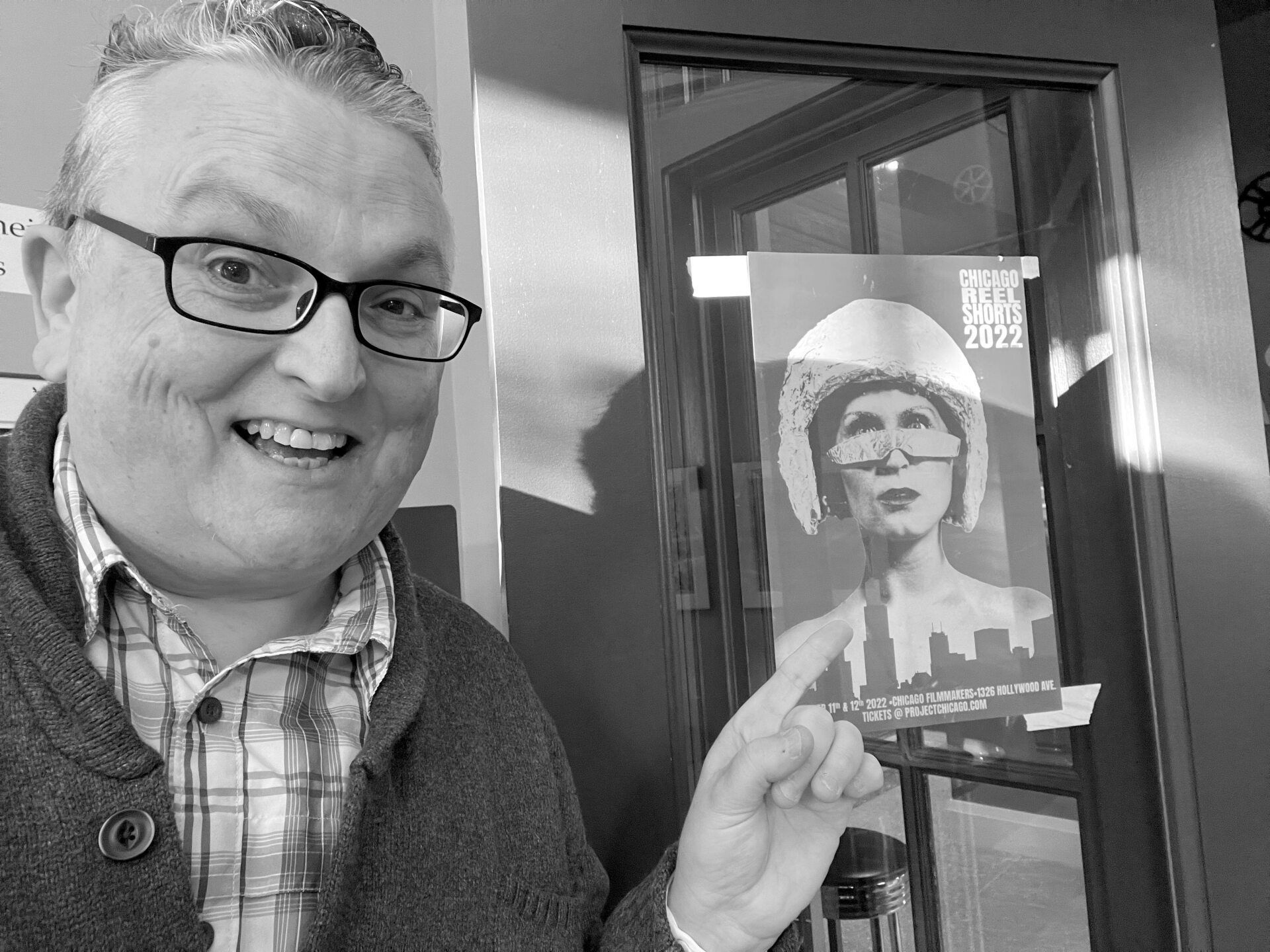Man smiles next to a poster. The poster has a woman with glasses. Black and white.