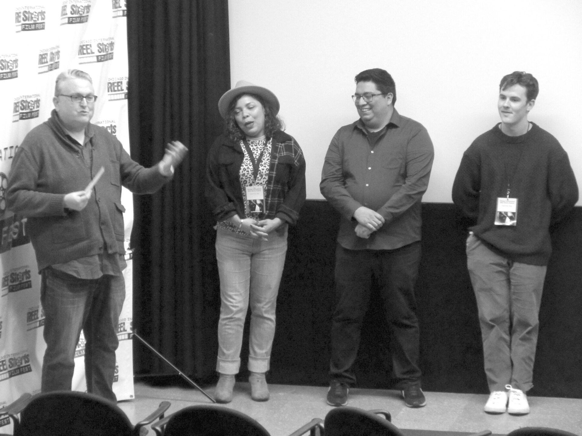 Four people standing on a stage in front of a banner. A man speaks into a microphone, three others listen.
