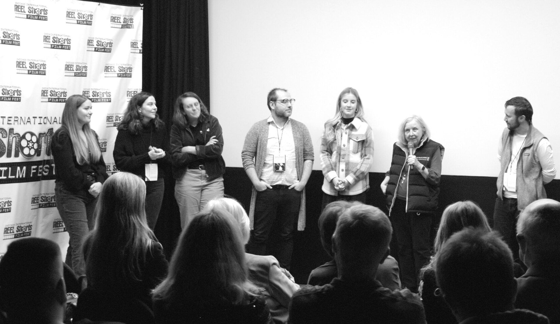 A panel of eight people stands on a stage. They are at a film festival with an audience in front of them.