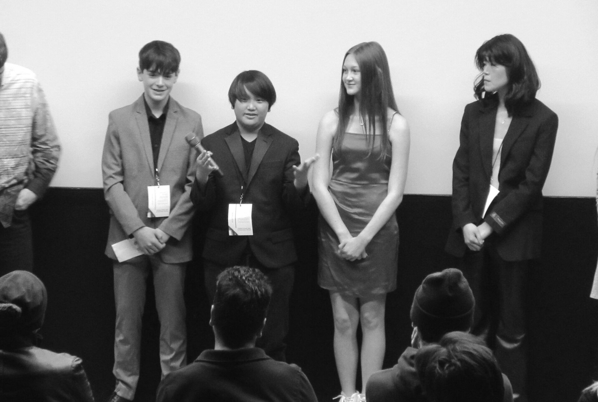 Four young people on stage with a microphone. One is speaking. Audience members are in the foreground.
