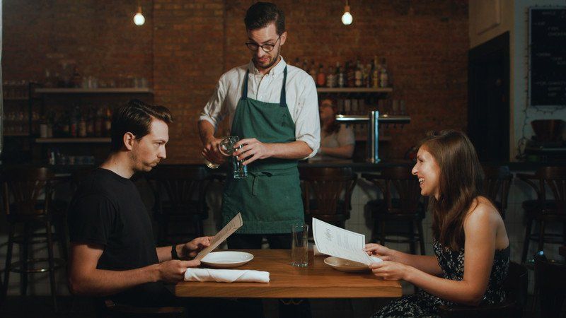 A waiter pours water for a couple seated at a restaurant table, brick wall backdrop.