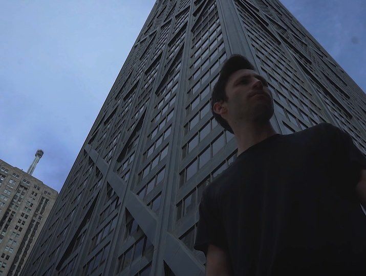 Man stands in front of a tall, dark skyscraper on a cloudy day.