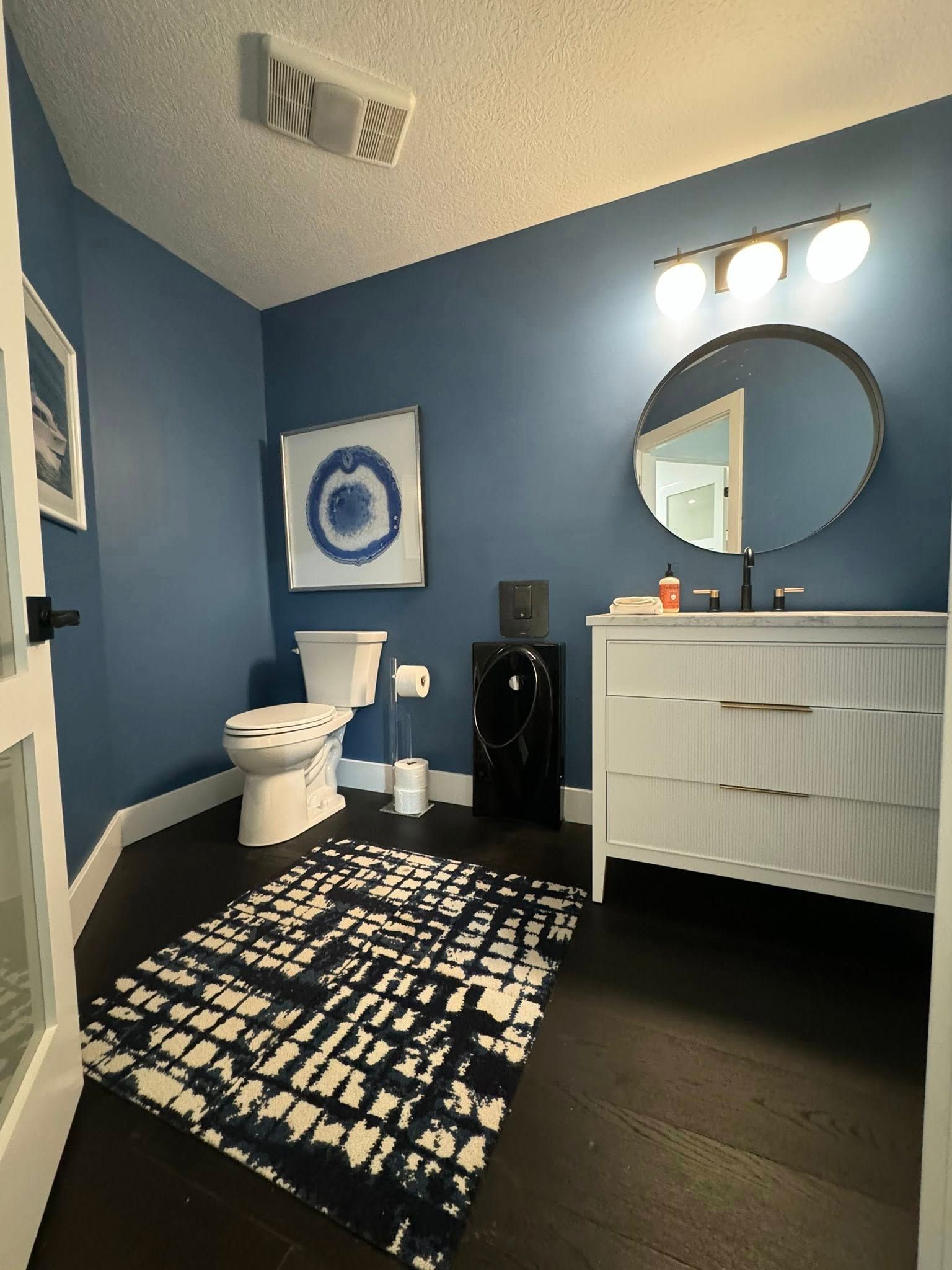 Blue bathroom with white vanity, round mirror, and patterned rug.