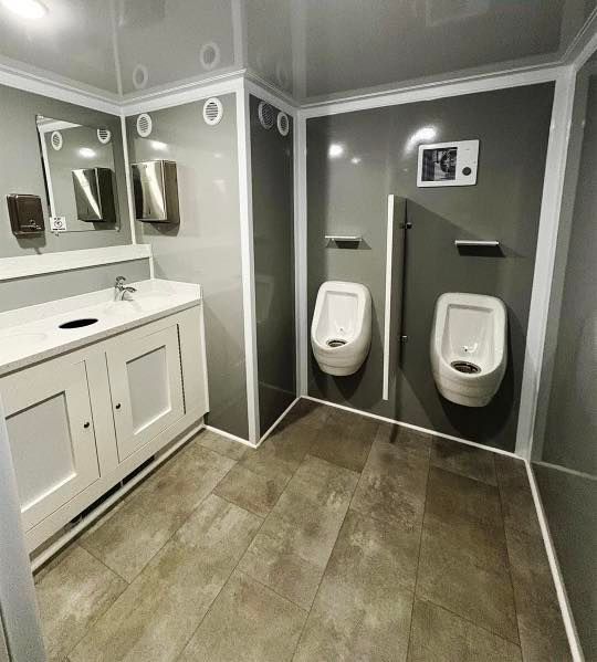 Accessible restroom with two wall-mounted urinals and a sink vanity with mirrors, in gray tile and walls
