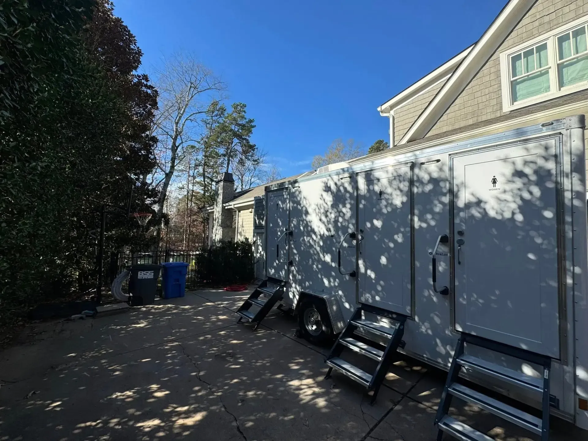 White storage containers along a driveway beside a house, with trees casting shadows on the wall.