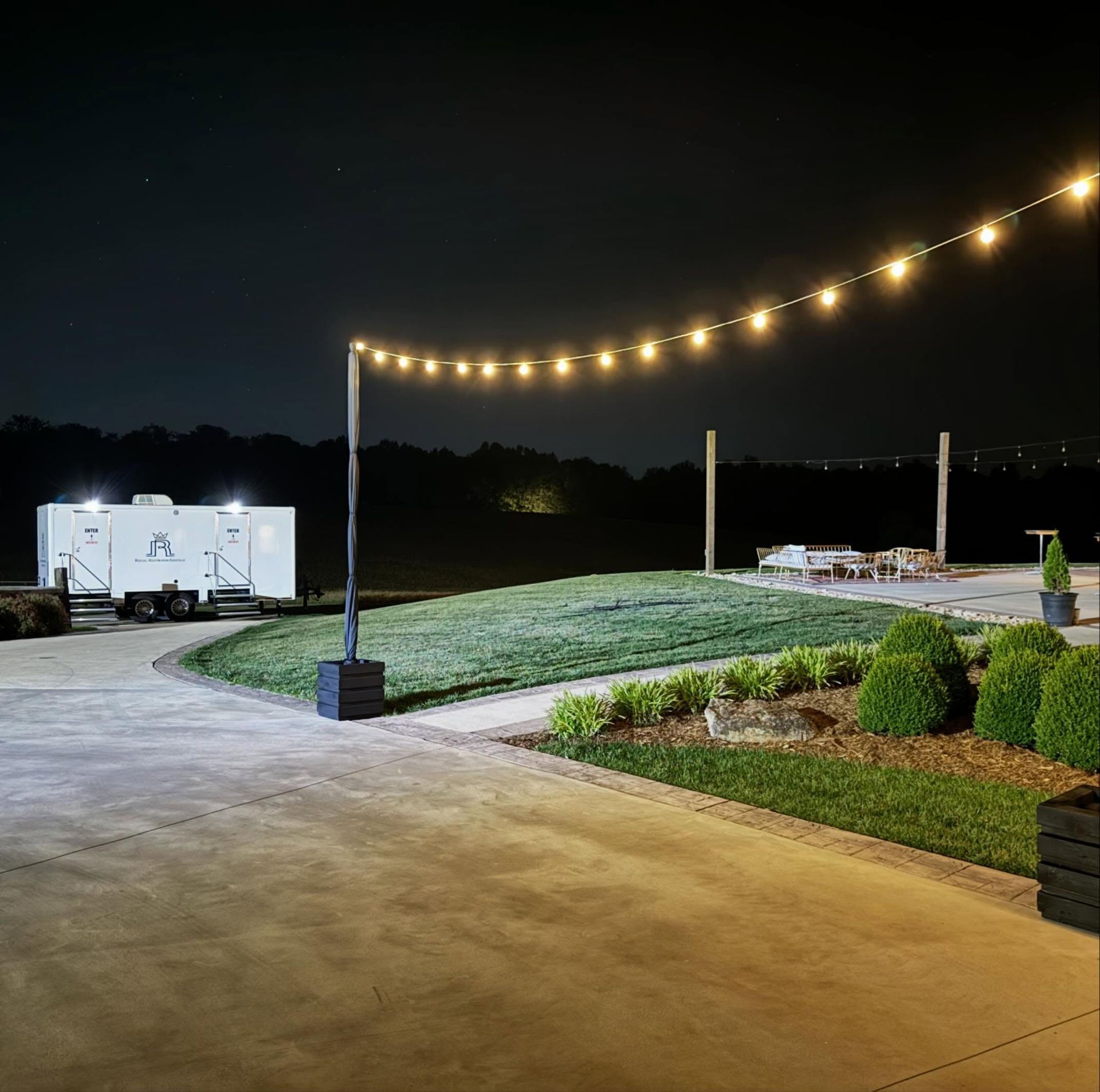 String lights over a paved patio at night beside a grassy yard and white camper trailers.