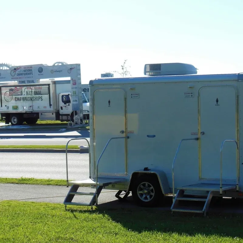 Blue trailer restroom unit parked on grass beside a road with a white truck in the background