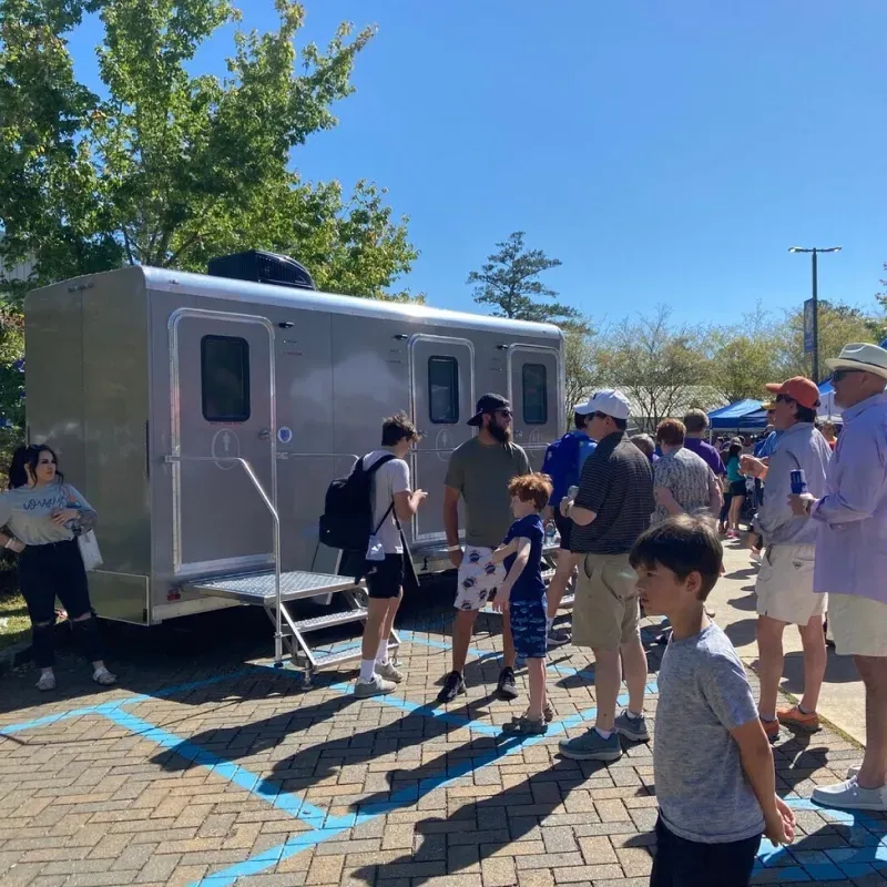People gathered near a silver trailer in a sunny outdoor park or plaza.
