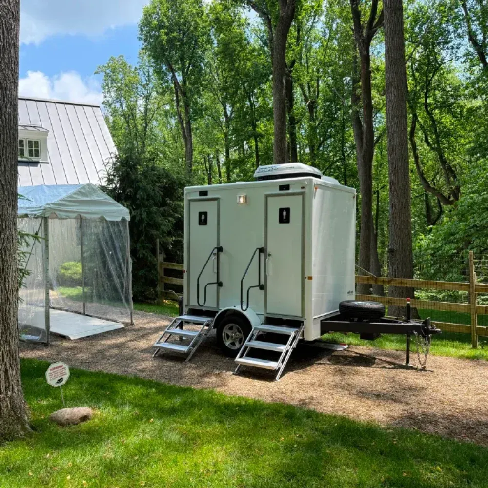 Two portable white restroom trailers with stairs in a wooded campsite beside a small building