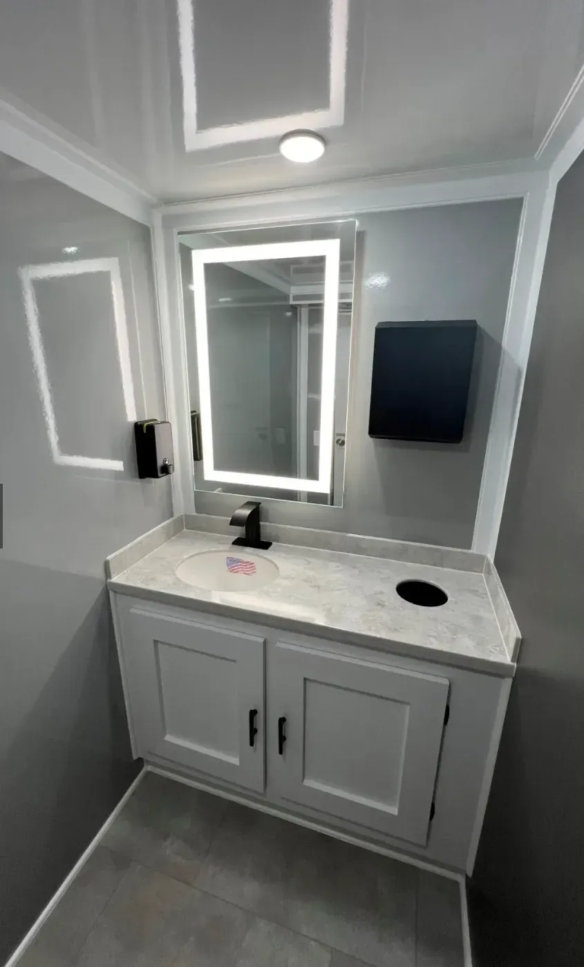Small gray bathroom vanity with mirror, sink, wall light, and mounted paper towel dispenser.