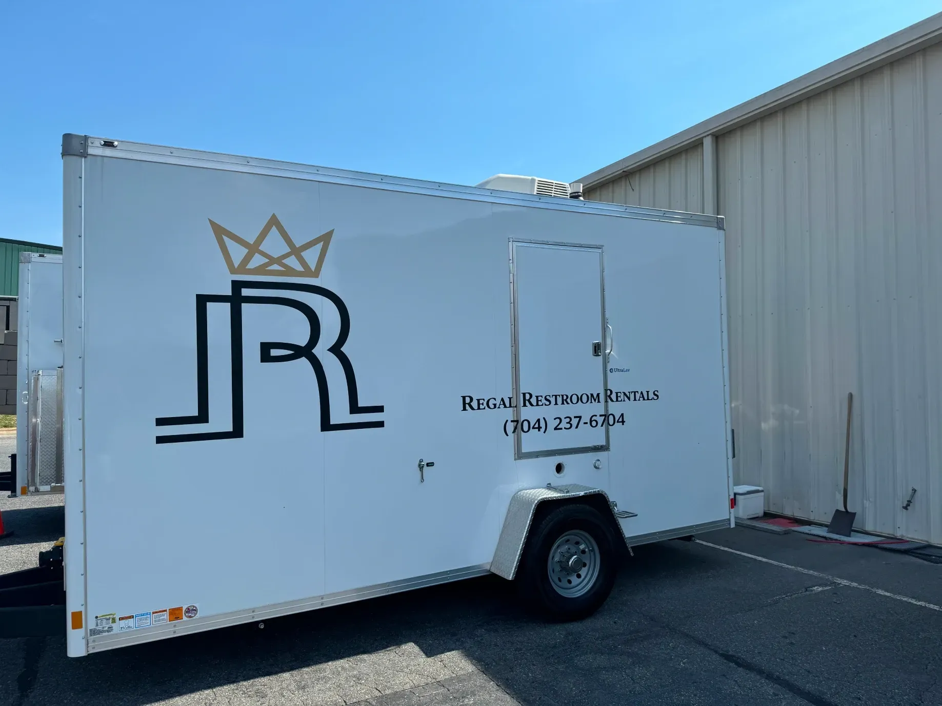 White enclosed trailer with a crown logo and stylized “JR” parked beside a metal building.