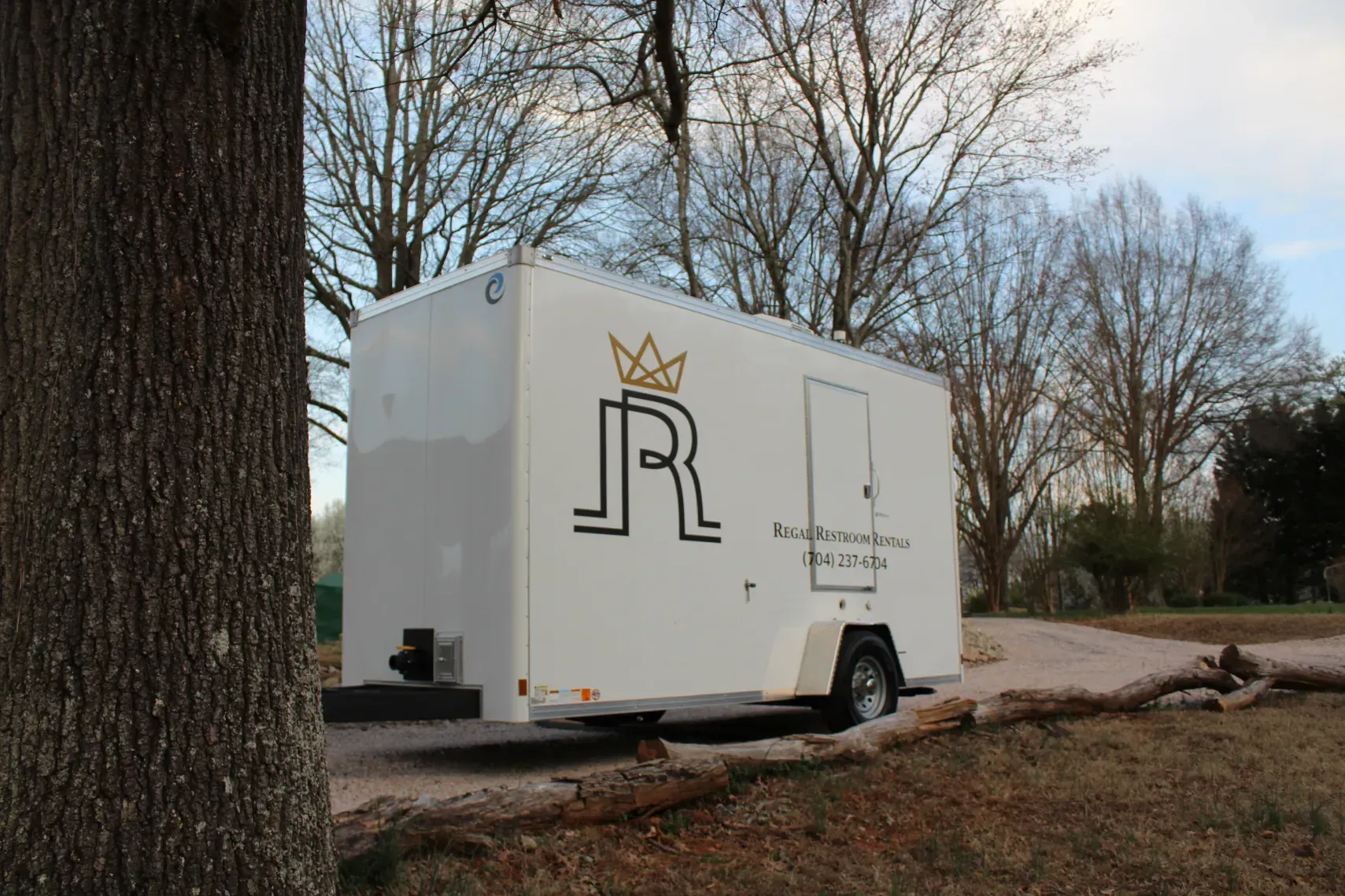 White trailer with a crowned logo parked beside a large tree on a quiet roadside