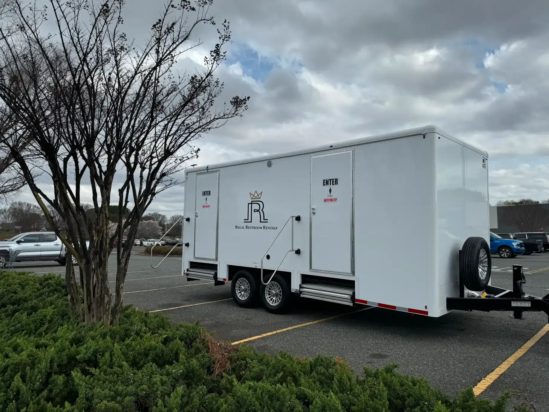 White enclosed cargo trailer parked in a lot beside a tree under cloudy skies.