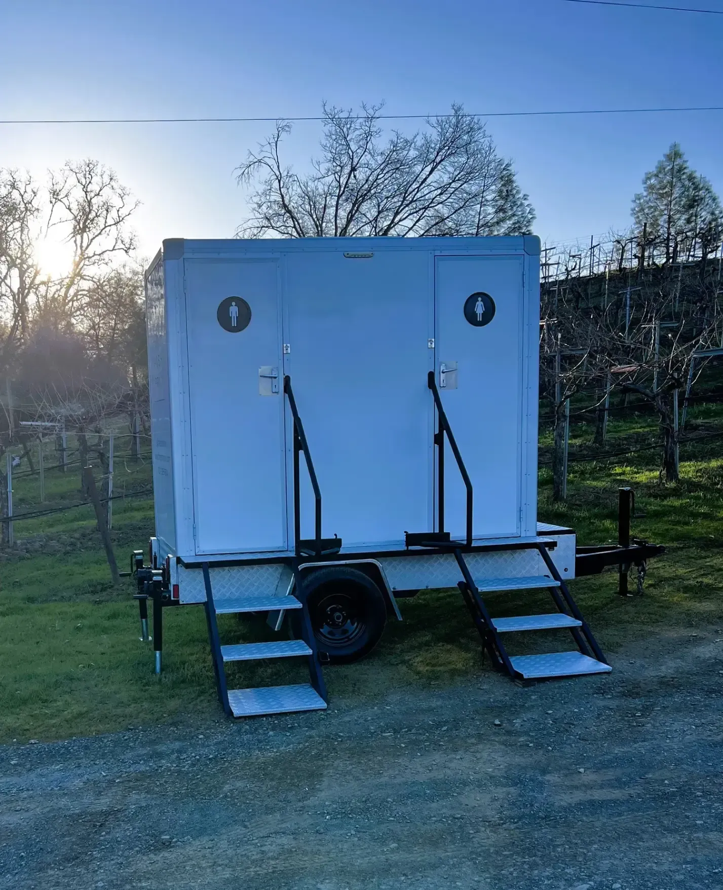 White portable restroom trailer with two doors and ramps, parked outdoors on grass at sunset.