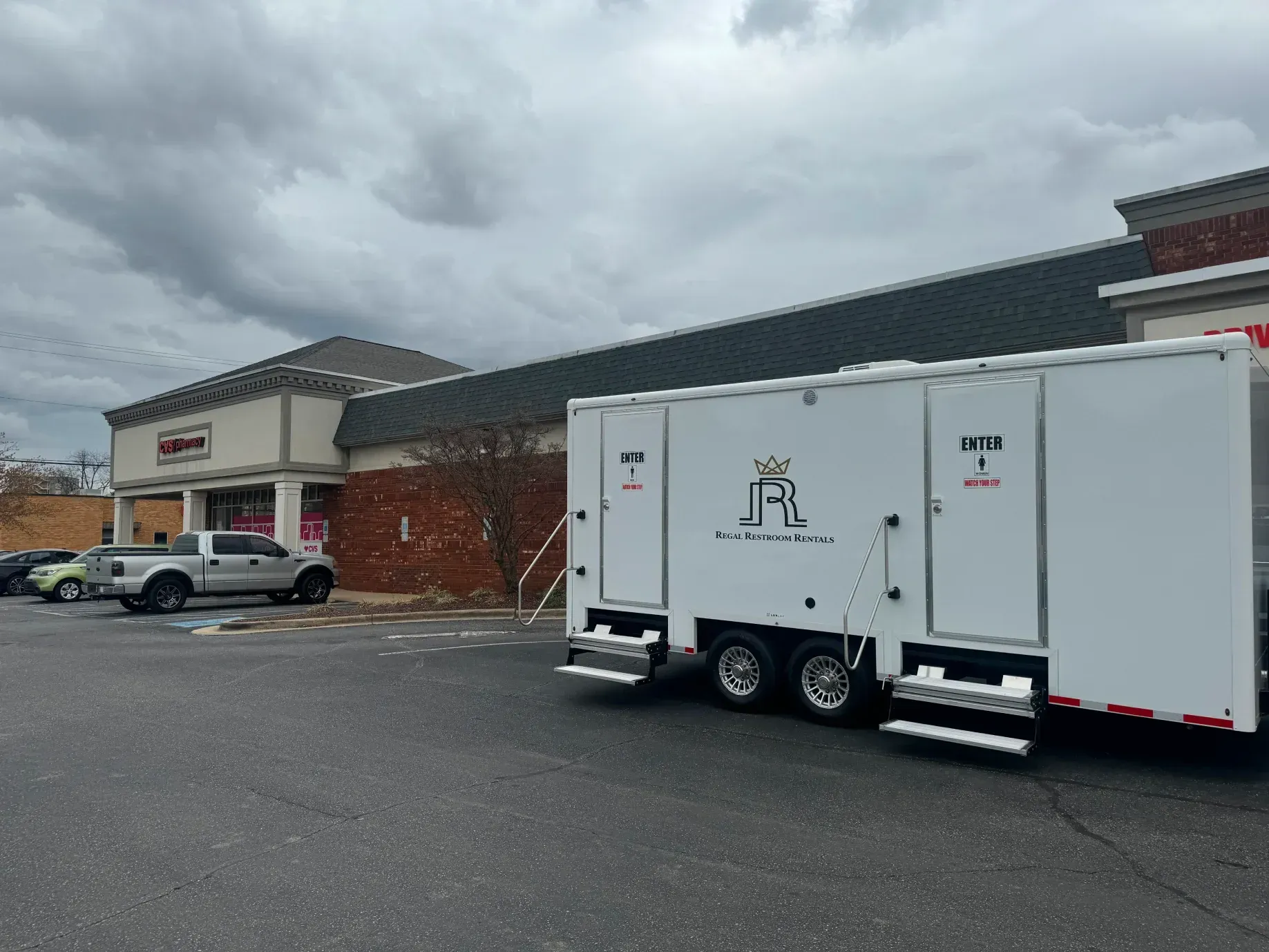White trailer parked in front of a brick shopping center under a cloudy sky.
