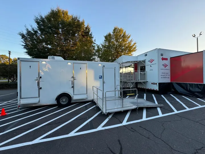 White mobile trailer and ramp in a parking lot beside a larger red-and-white trailer at sunset