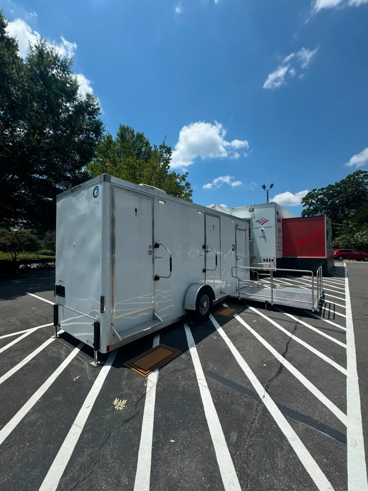 White utility trailer parked in a marked lot under a bright blue sky, with a red truck nearby.