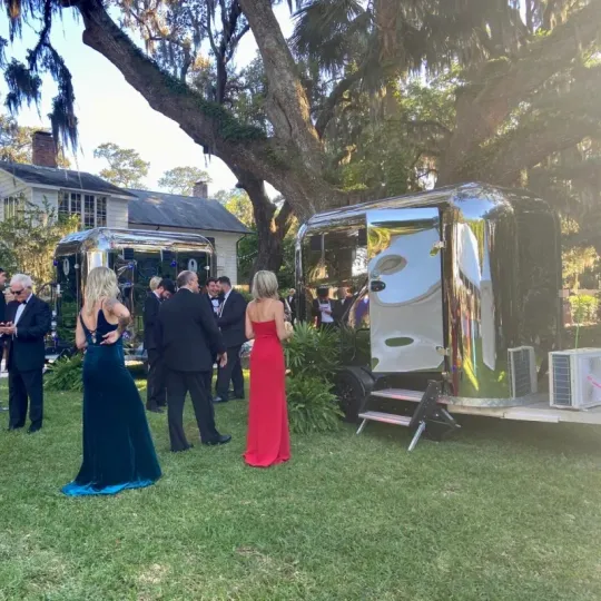 Guests stand near silver trailers in a grassy yard under a large tree at an outdoor event.