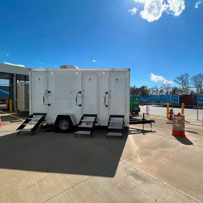White portable restroom trailer with ramps in a sunny parking lot, near orange cones and a construction fence