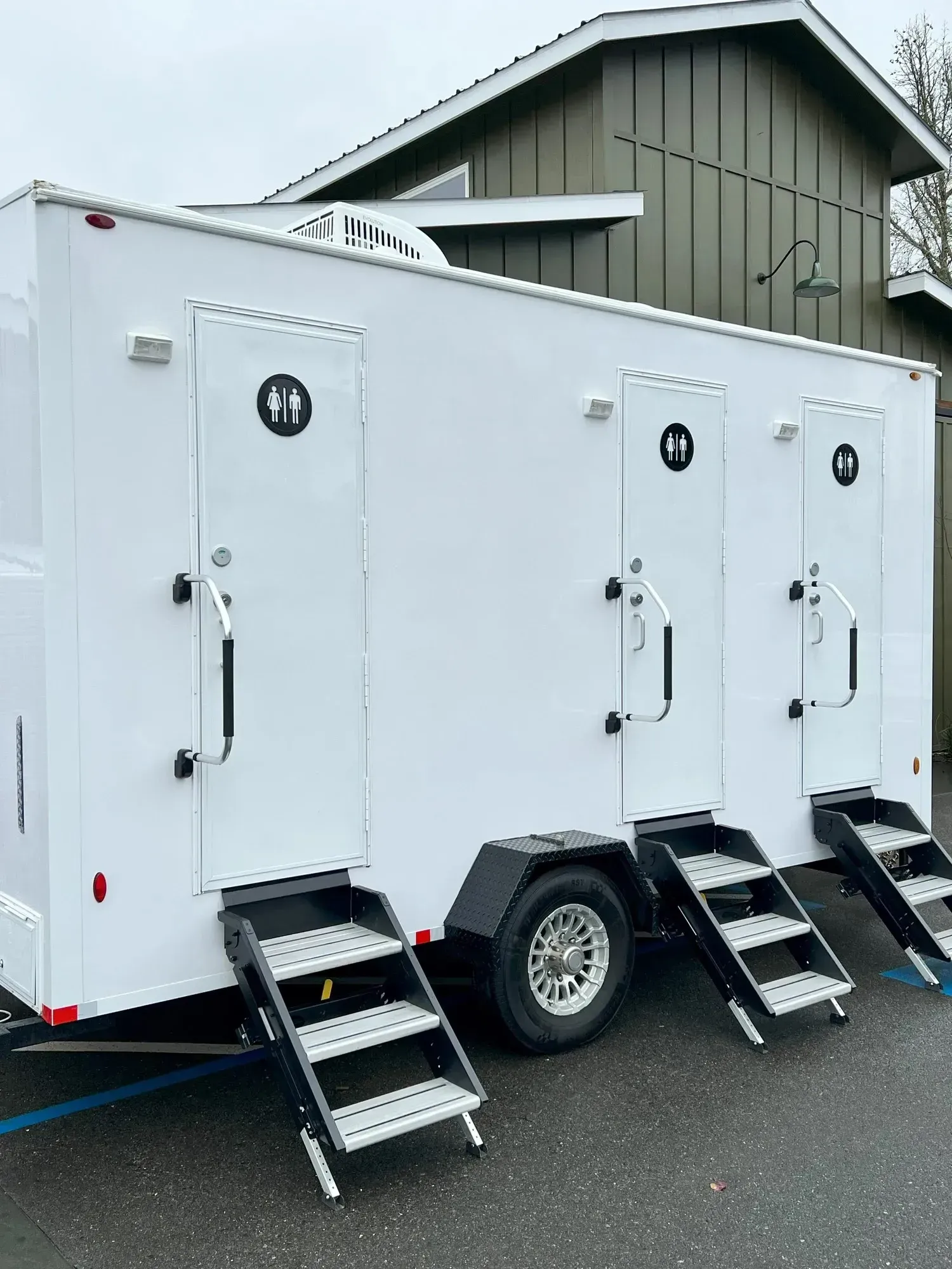 White portable restroom trailer with three doors and stairs, parked outside a building.