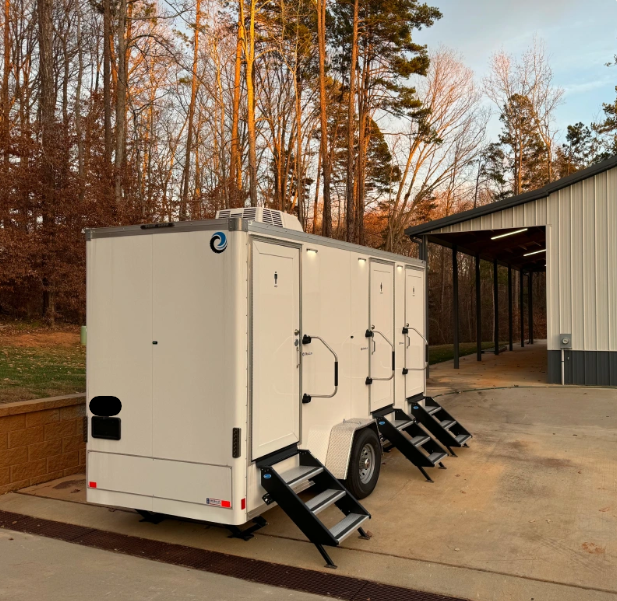 White mobile trailer units with black stairs parked on a concrete lot beside a building, with trees in the background