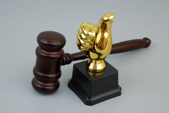 Wooden gavel next to a gold thumbs-up trophy on a black base, against a gray background.