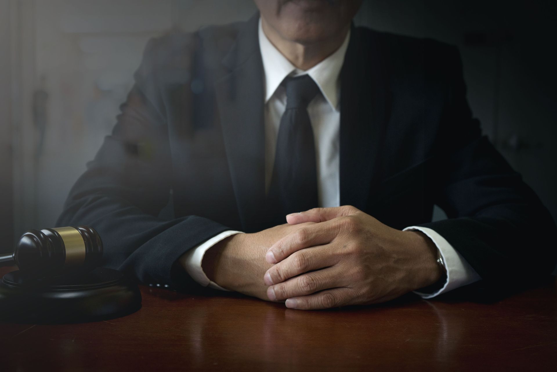 Judge at desk, hands clasped, gavel in view. Dark suit, dim setting.