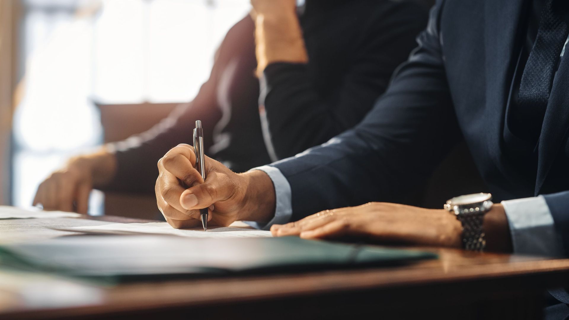 Person in suit signing document at a table, with another person partially visible.