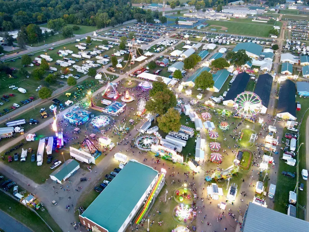 Aerial view of a fair with rides, food stands, and crowds at night; lights and structures are visible.