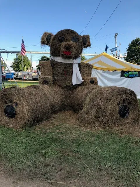 Large teddy bear sculpture made of hay with a white scarf at an outdoor fair.