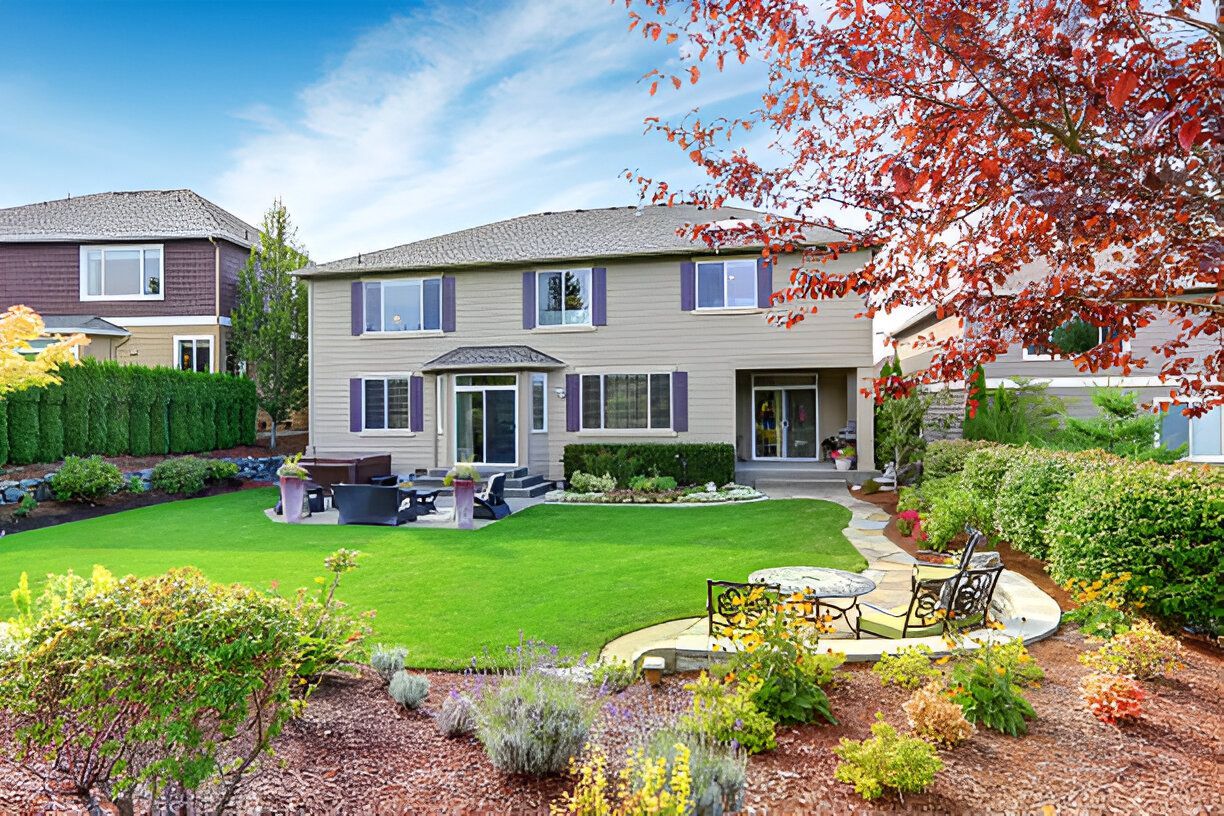 Two-story beige house with green lawn, stone patio, and red-leafed tree; blue sky background.
