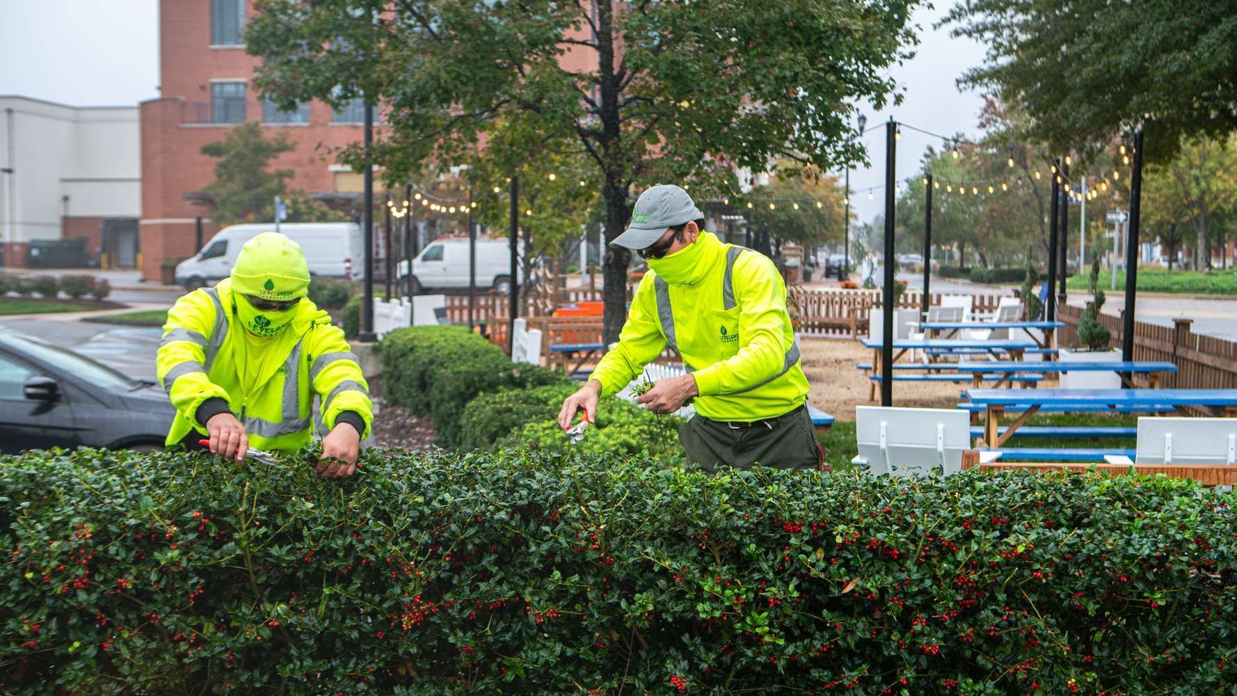 Two people in neon yellow jackets trimming a green hedge outdoors near a seating area.