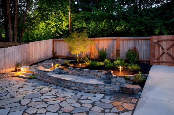 Backyard patio with stone paving, low wall, and illuminated landscaping against a wooden fence.