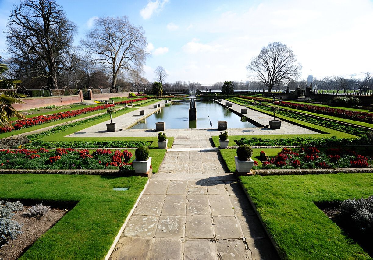 Formal garden with a rectangular pond, surrounded by green lawns, flower beds, and trees under a blue sky.