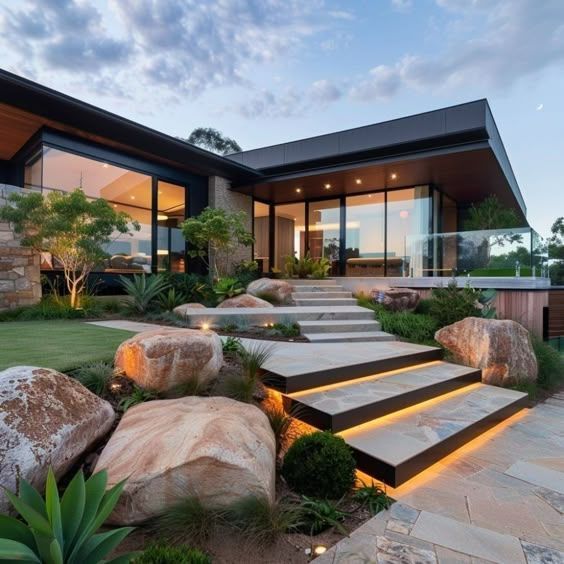Modern house with illuminated steps, large windows, and landscaped garden with boulders.