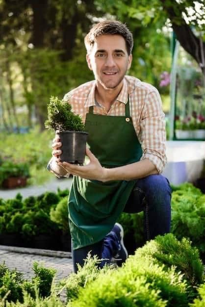 Man kneeling in a garden, holding a potted plant. Wearing apron, checkered shirt, smiling.