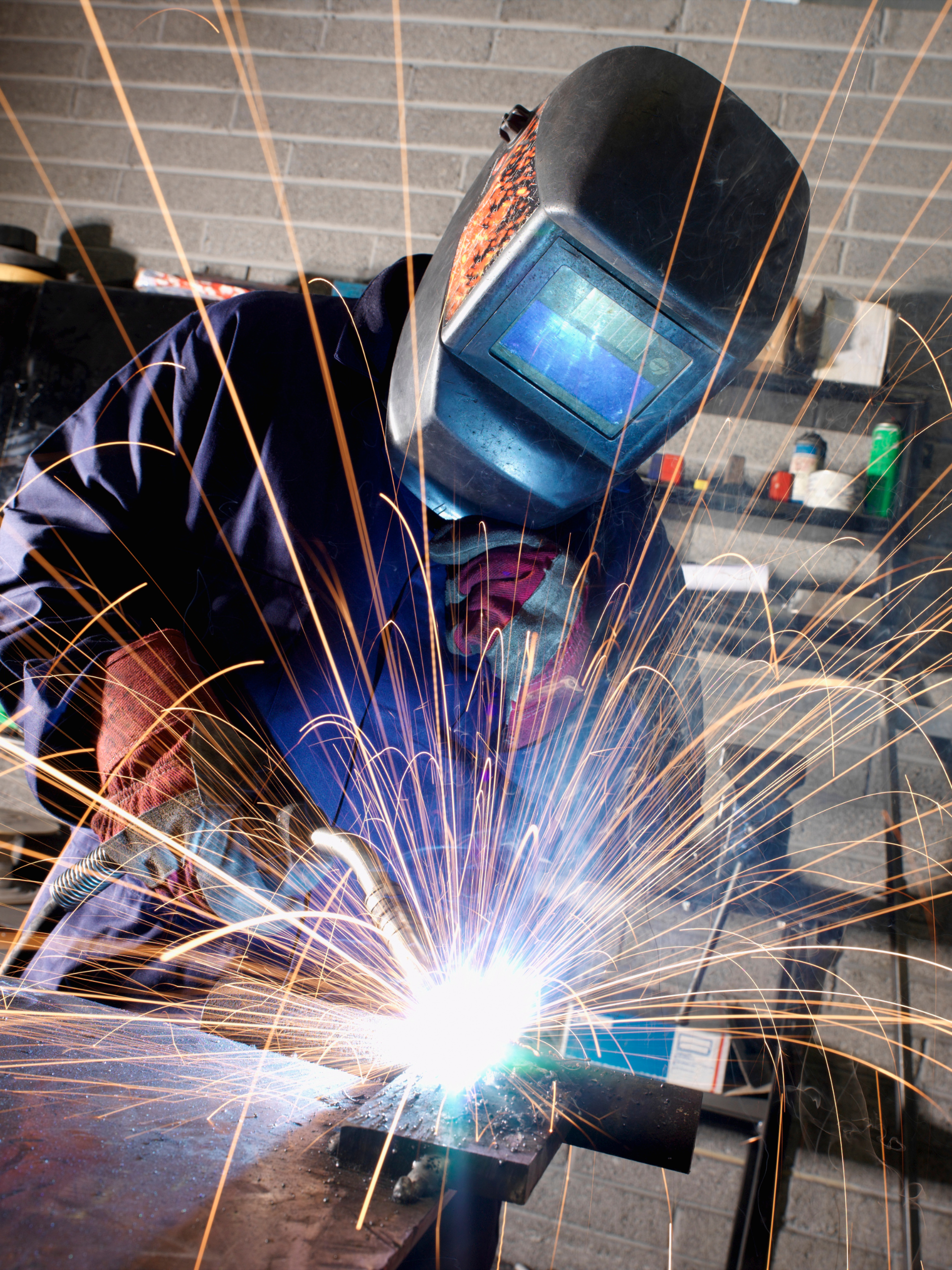 Welder wearing a welding mask while he welds a piece of metal with spark flying off