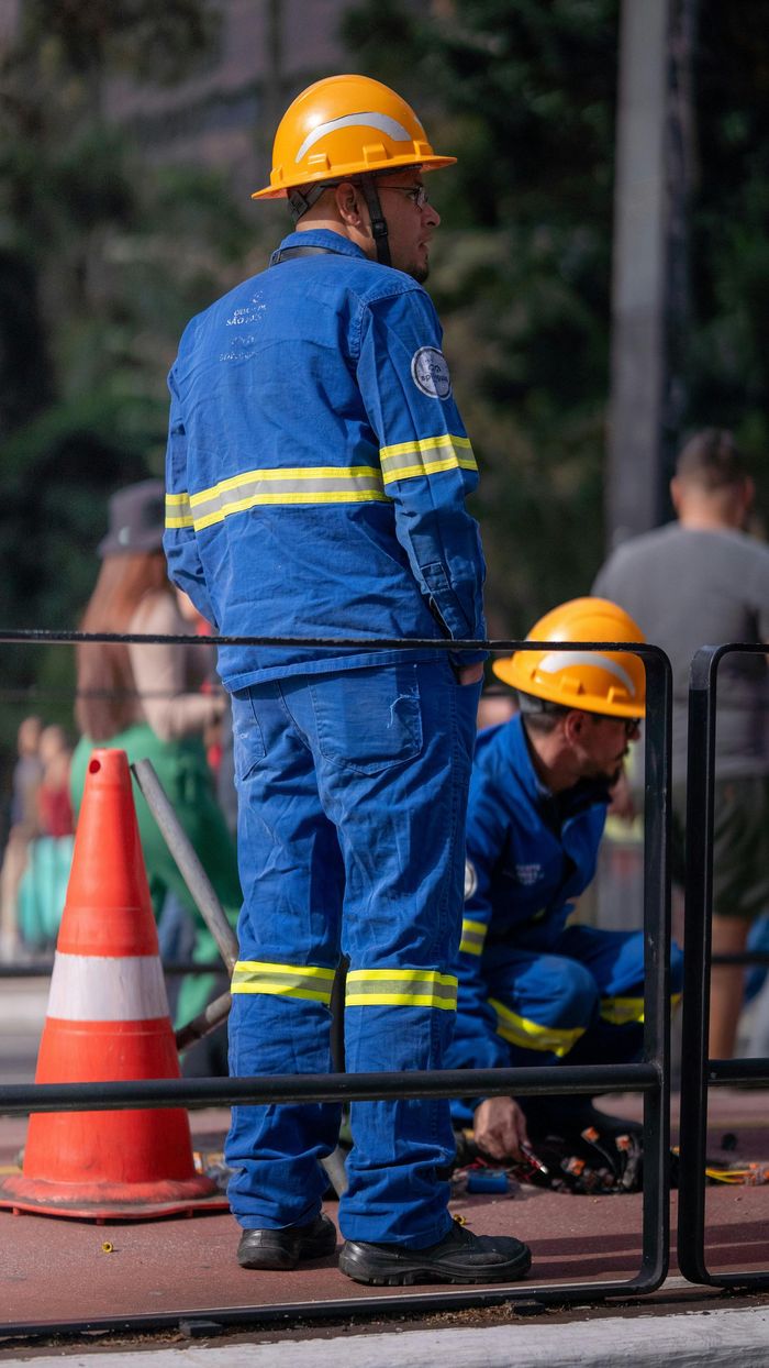 A man in a blue jumpsuit and a yellow hard hat is standing next to a fence.