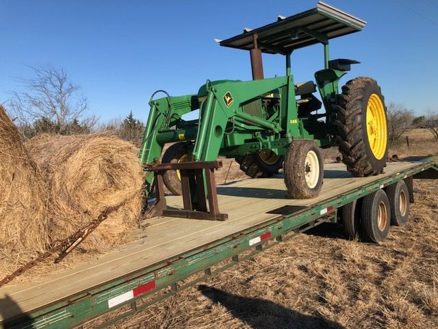 Green John Deere tractor with loader on a flatbed trailer, next to hay bales.