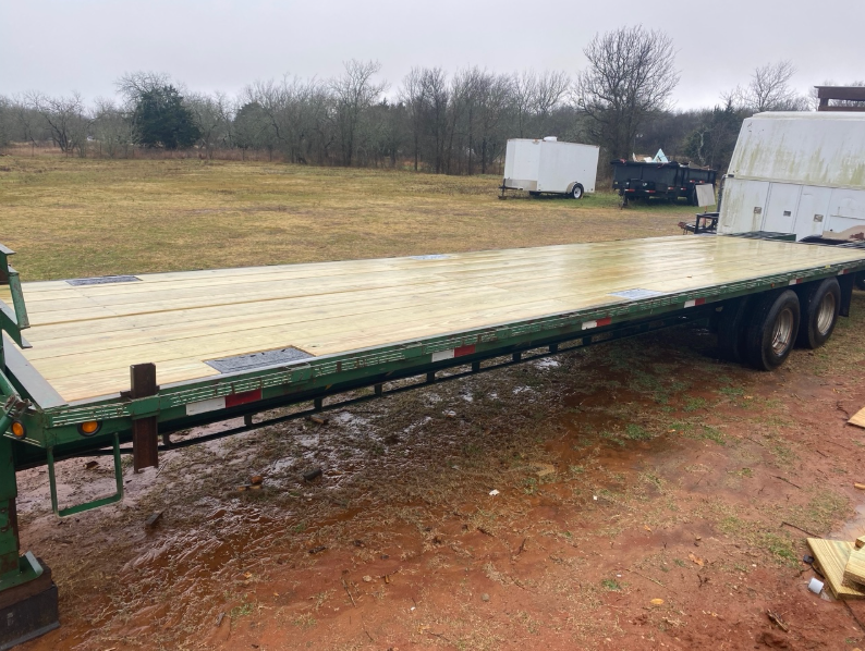 Flatbed trailer on a muddy field, trees and buildings in the background. Green and brown colors dominate.