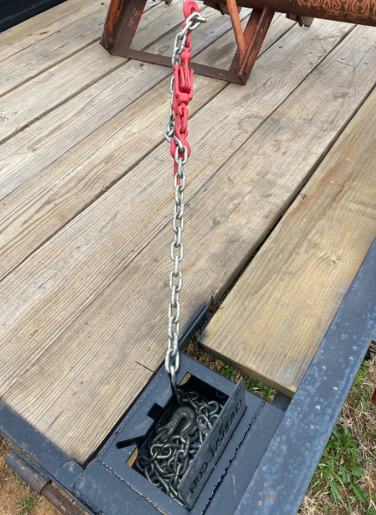 A chain hangs from a red hook on a wooden trailer bed, leading into a storage compartment.