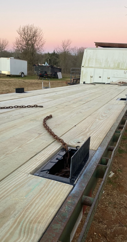 A flatbed trailer with a wooden deck and recessed chain tie-downs, parked in an open field at sunset.