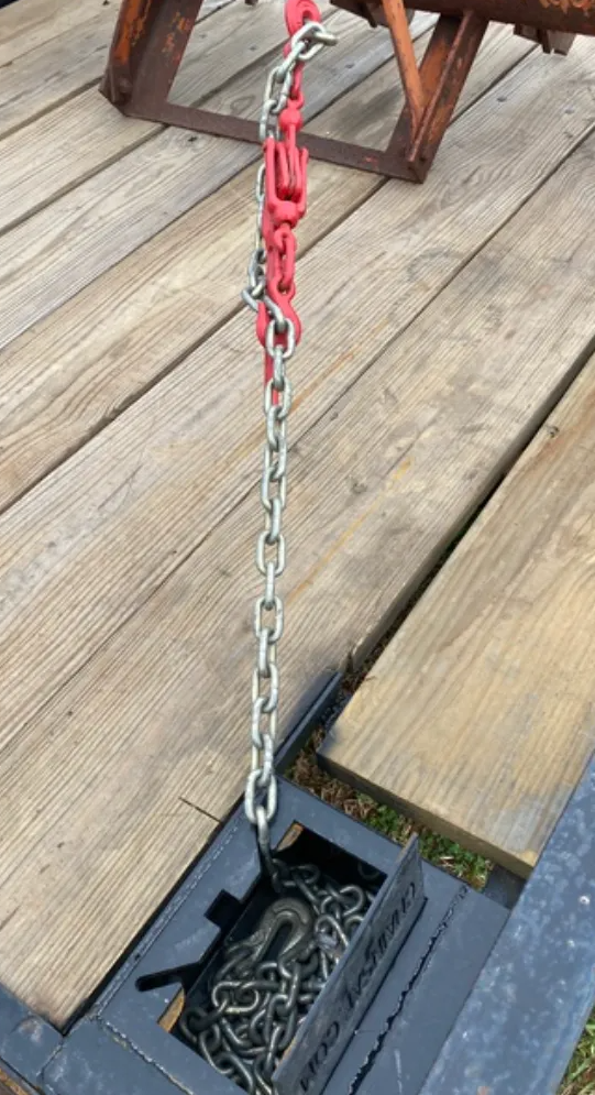 A red chain binder attached to a metal chain, which is anchored in a recessed storage box on a wooden trailer deck.
